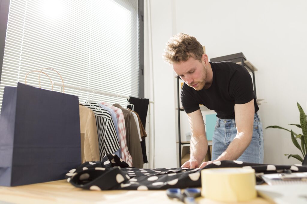A person stands at a table, focused on folding a black polka-dot garment, with shopping bags and plants in a bright room.
