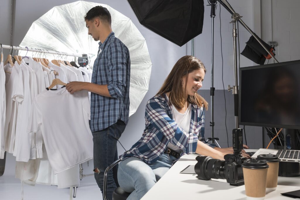 A photoshoot setup featuring a man reviewing clothing on a rack and a woman working on a computer with photography equipment nearby.