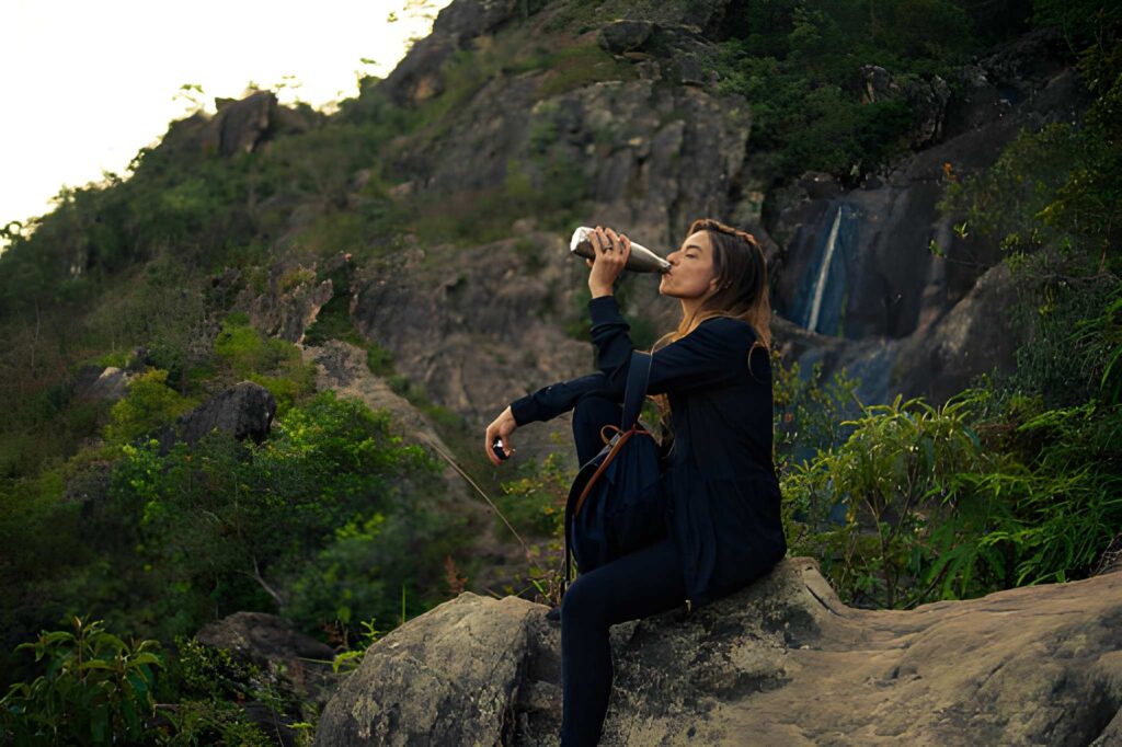 A woman sitting on a rock in a serene outdoor setting, sipping from a bottle, with a waterfall and lush greenery in the background.