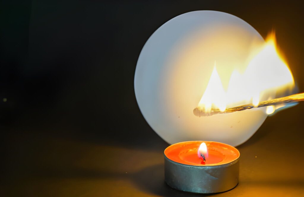 A match ignites above a lit candle, with a blurred white circle in the background, creating a striking contrast of light and shadow.