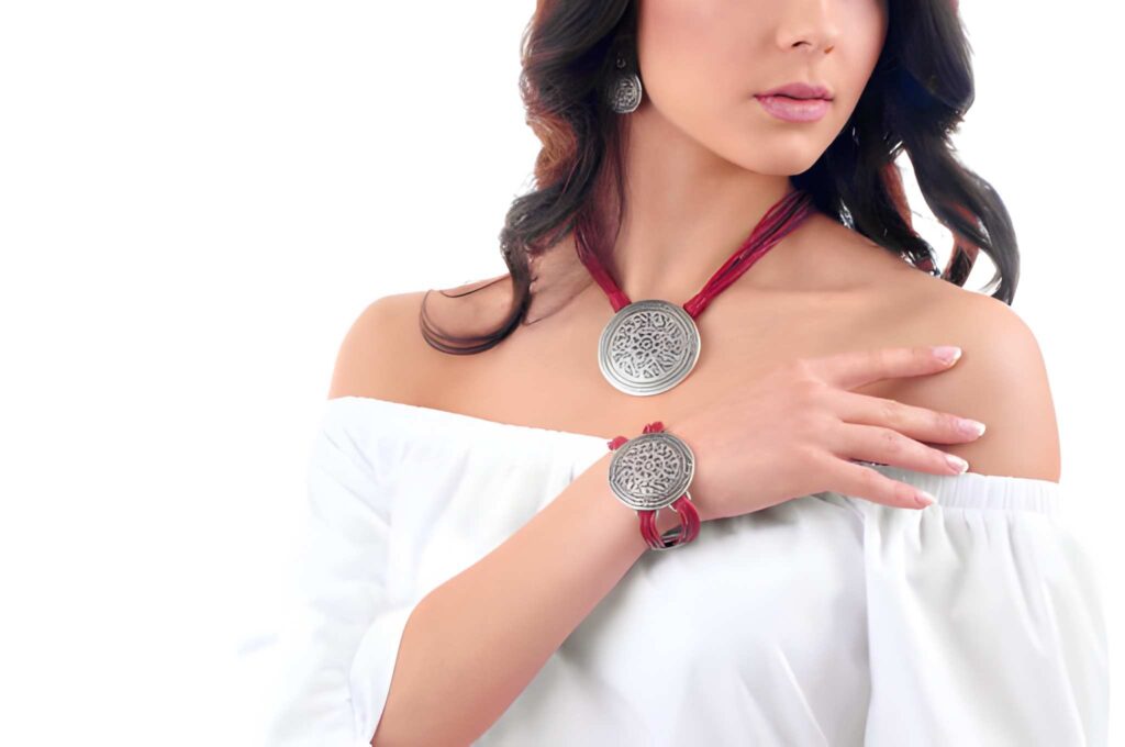 A woman models silver jewelry, wearing a large pendant necklace and bracelet, with long, wavy hair and a white off-shoulder top.