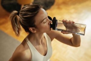 Person in a white tank top holding a clear water bottle over a wooden floor.