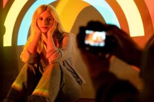 Person posing with colorful light arcs in the background, being photographed.