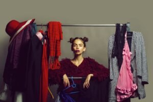 A woman in a seated position, looking at a rack filled with clothes, contemplating her choices among the displayed apparel and posing for eCommerce fashion photography.