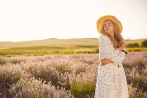 A woman wearing a polka dot dress and hat stands gracefully amidst a vibrant lavender field, surrounded by blooming flowers.