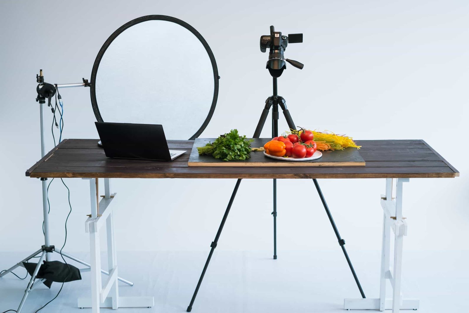 Tabletop photography setup with camera, lighting equipment, laptop, and fresh vegetables.