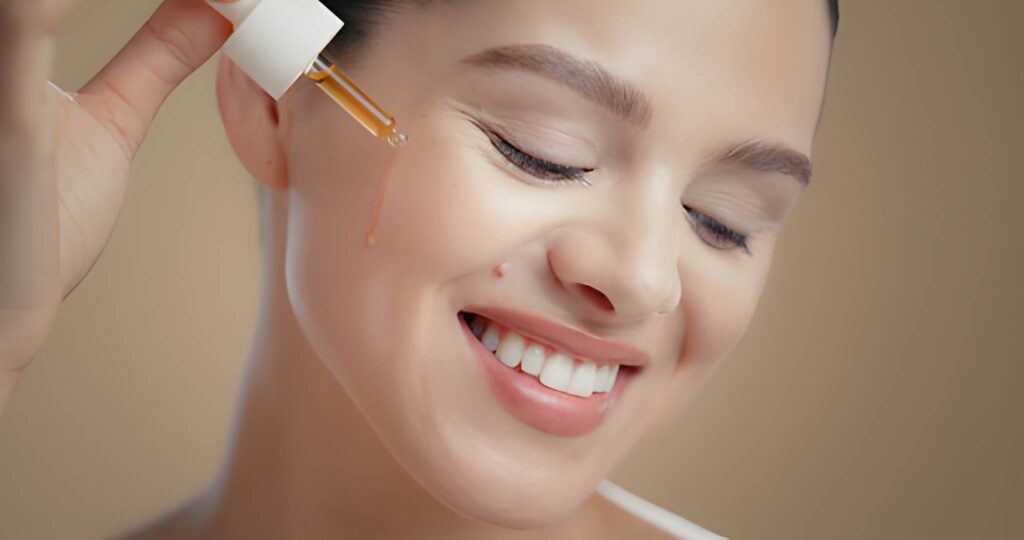 Smiling woman with eyes closed receiving a drop of amber-colored facial serum applied near her cheekbone with a dropper.
