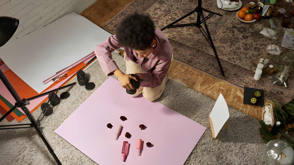 Overhead view of a person photographing makeup on a pink background. Surrounding area includes props and studio equipment, conveying a creative, focused atmosphere.
