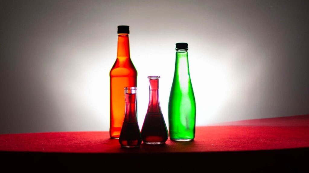 Three decorative bottles in red, green, and purple glass are silhouetted against a bright background on a red surface.