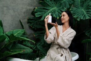 a woman in a bathrobe holding a bottle of cosmetic product with plants in the background.