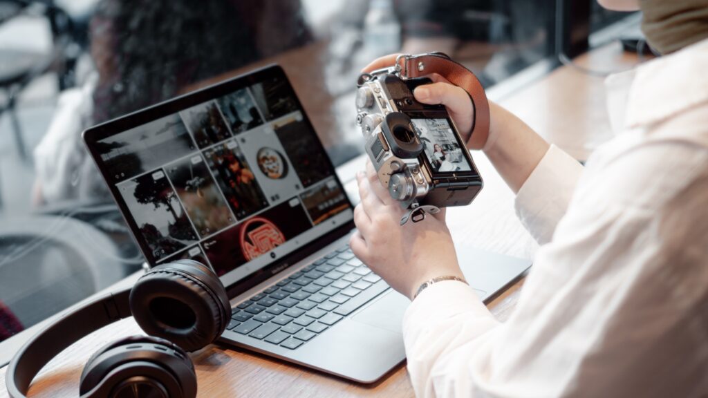 A person in a white shirt holds a vintage camera beside a laptop displaying photos. A pair of headphones rests nearby on the wooden table.