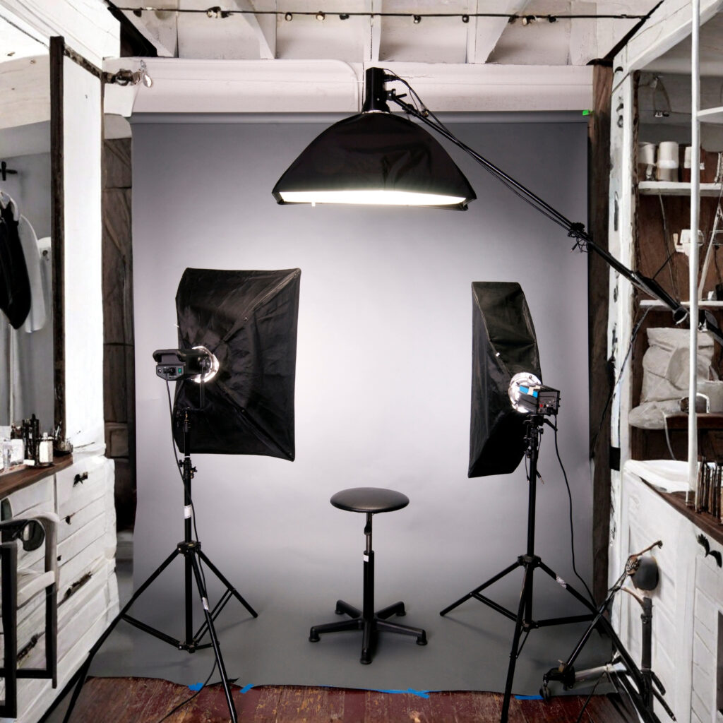 A photography setup featuring a gray backdrop, two softbox lights, a camera on tripods, and a black stool in the center.