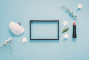 Flat lay of a perfume bottle, lipstick, empty frame, and flowers on a blue background.