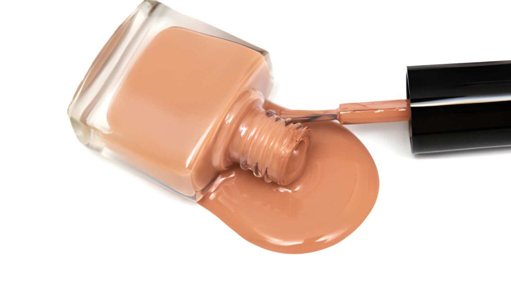 Clear glass bottle of nude or beige nail polish spilled on a white surface with the applicator brush resting beside it.