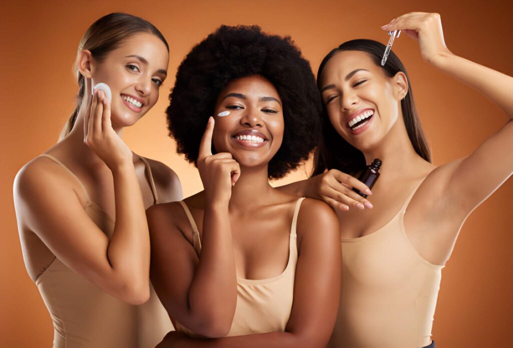 Three smiling women of diverse ethnicities apply skincare products against a warm brown background.