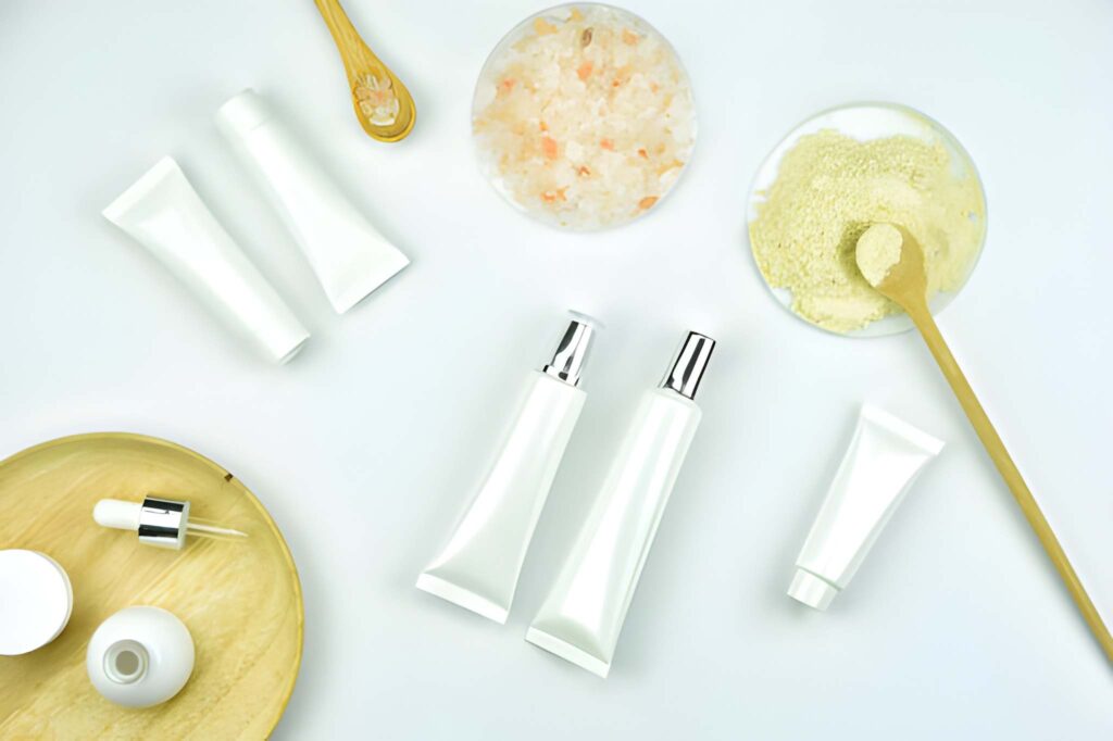 White cosmetic tubes, wooden spa tools, and containers of pink and beige scrubs are arranged artistically on a white background, conveying a minimalist spa theme.