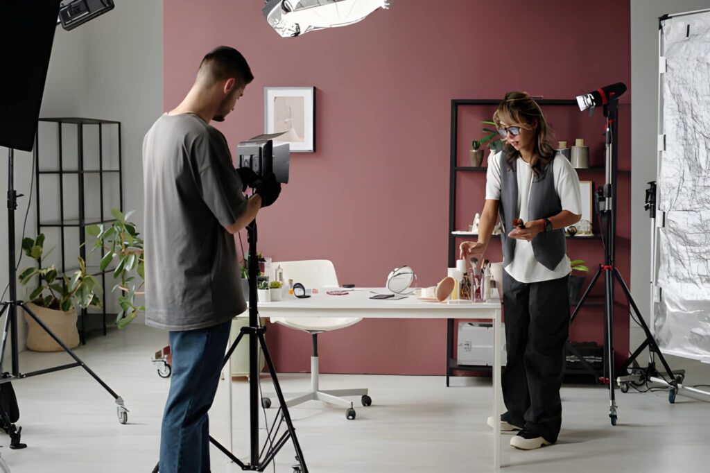 A photographer on a tripod adjusts a camera while a woman poses near a white table with products in a brightly lit studio setting with mauve walls.