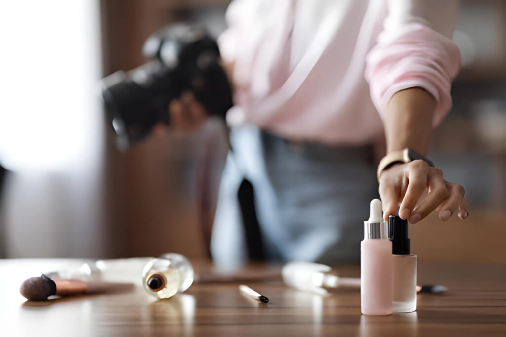 Person in a light pink shirt holding a DSLR camera while placing a small bottle of cosmetic product onto a table with other makeup items.