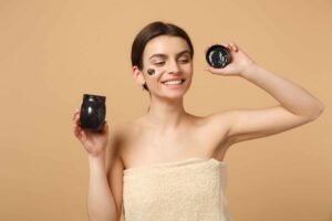 Person in a towel holds an open jar of cream with a beige background.