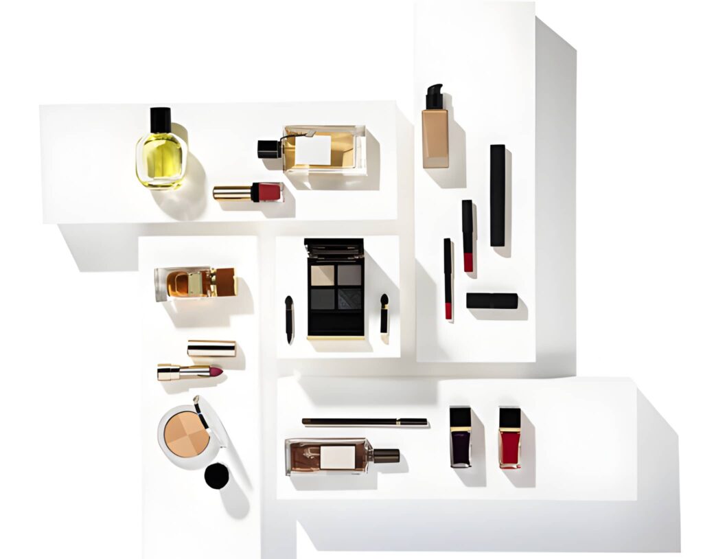 Assortment of luxury cosmetics, including perfumes, eyeshadow palettes, lipsticks, and foundation bottles, elegantly displayed on staggered white shelving against a bright white background.