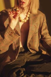 Woman in a houndstooth blazer holding glasses, with a large pearl necklace. Warm lighting.