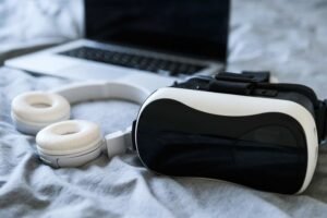 A VR headset and white headphones beside an open laptop on a bed.