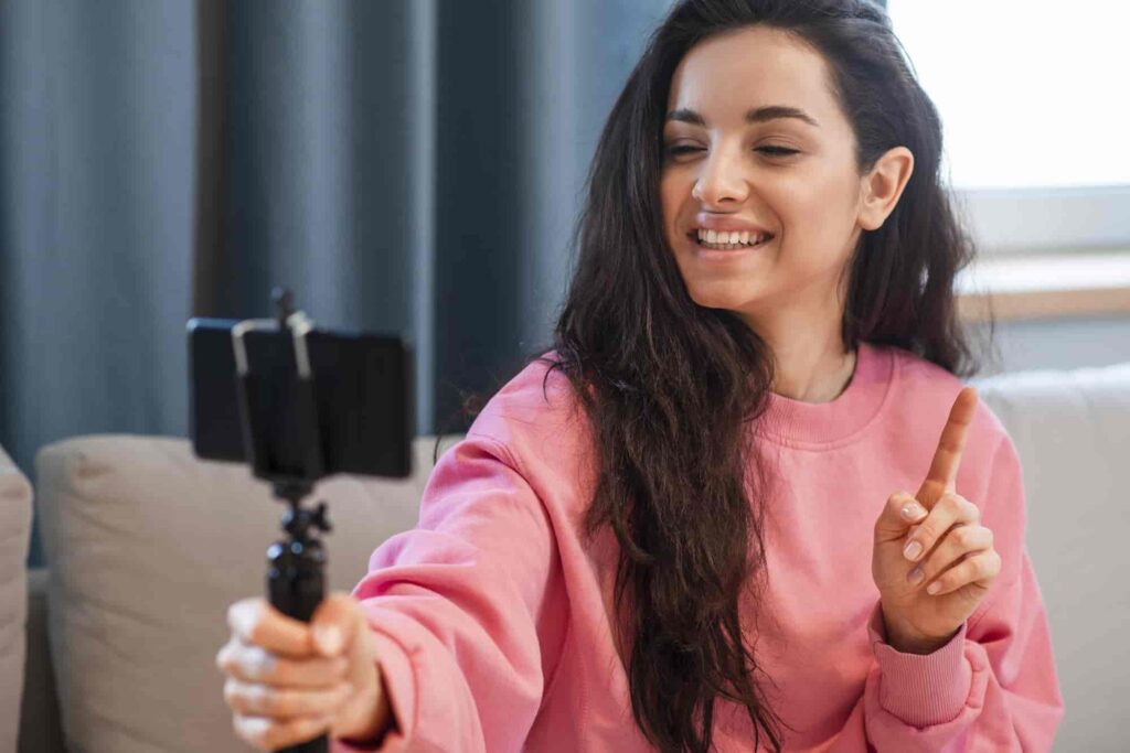 Creator recording short Instagram video clip on smartphone while sitting on a couch in casual pink sweatshirt.