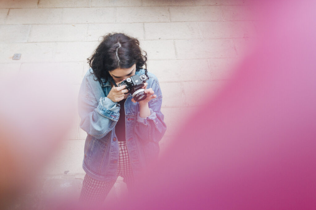 Content creator filming short Instagram video clip outdoors with camera, wearing denim jacket and checkered pants.