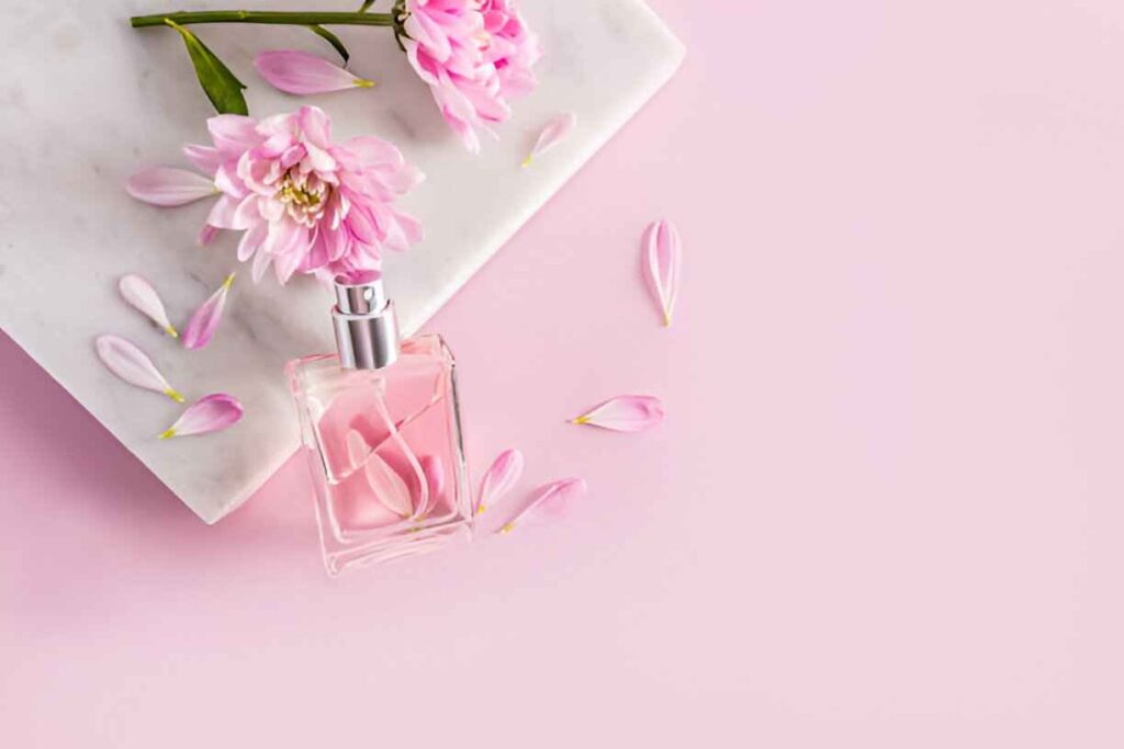 A pink floral perfume bottle rests on a marble slab, surrounded by delicate pink petals and a pink background.