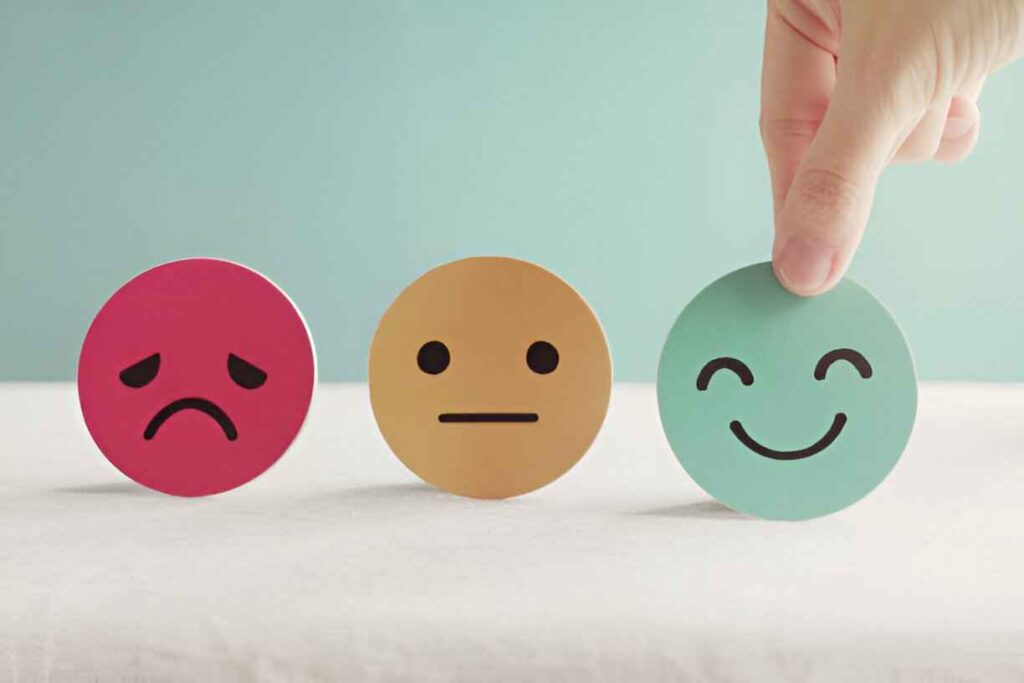 A hand lifts a smiling blue emoji card while two other cards display a sad red emoji and a neutral yellow emoji, symbolizing emotions.