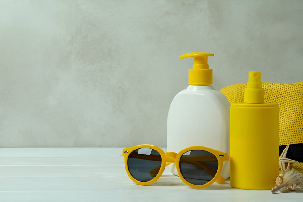 A white lotion bottle, a yellow spray bottle, and yellow sunglasses are arranged on a white surface with a textured beige background.