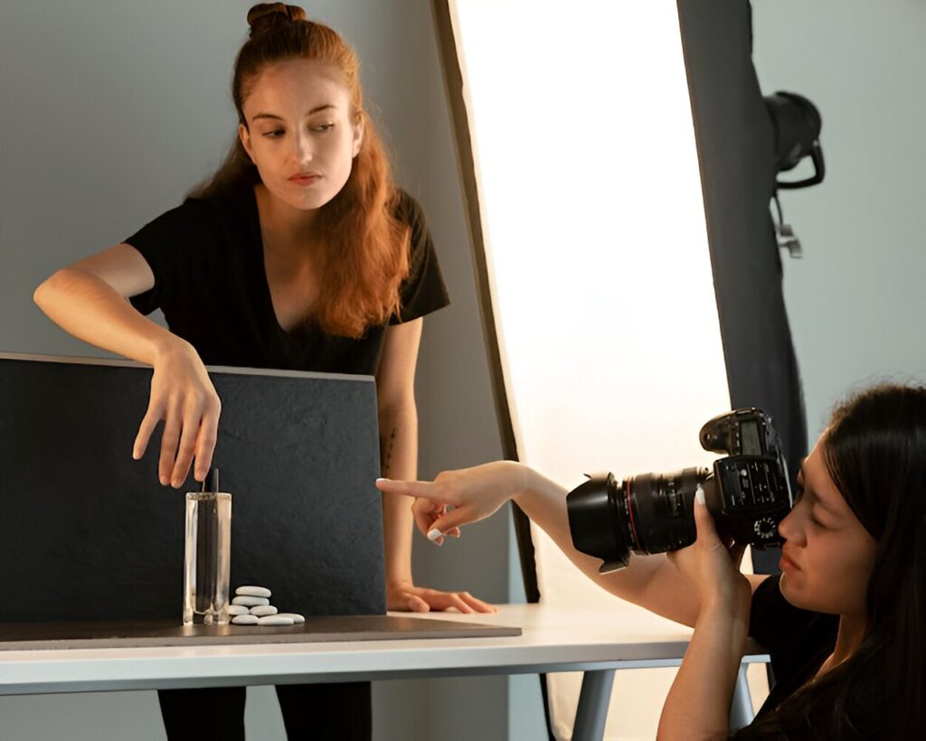 A photographer directs a model as she interacts with a glass and white pebbles on a shoot set, with professional lighting equipment present.