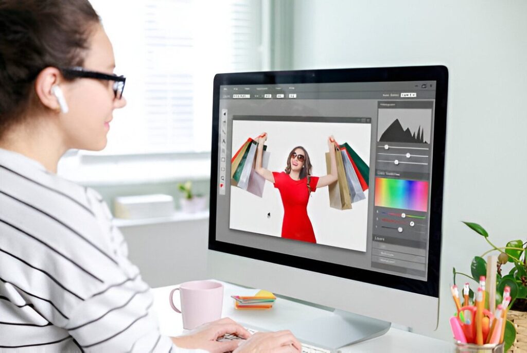 A person wearing headphones edits a photo of a woman in a red dress holding shopping bags on a computer screen. A plant and stationery are nearby.