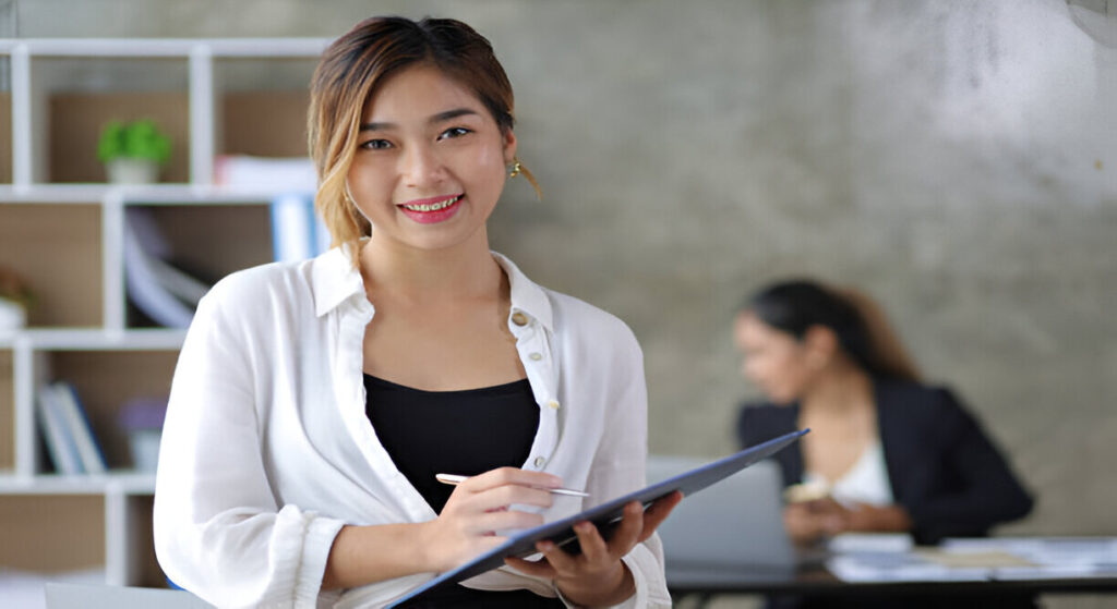 A woman in a white blouse holds a clipboard, focused on her work, with a modern office setting and a colleague in the background.