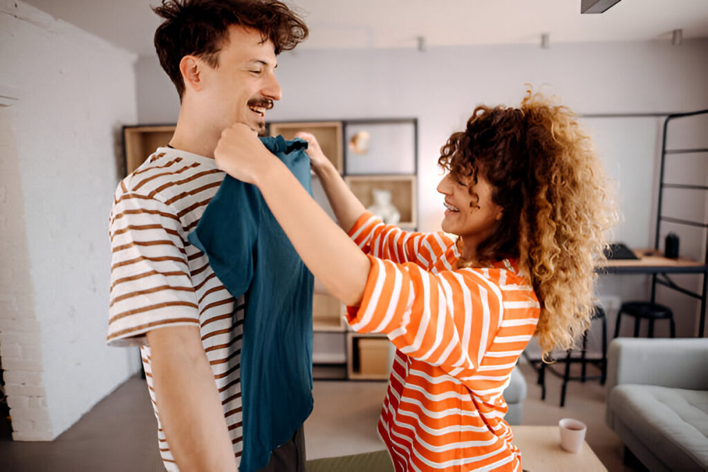 A person in a striped shirt assists another wearing a blue shirt, preparing for an outfit change in a cozy, modern room.