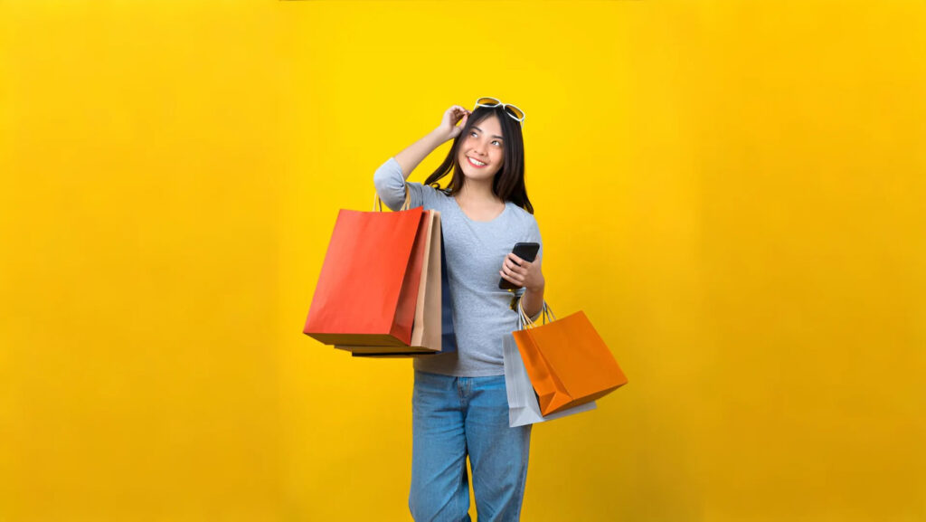 A person in a gray shirt and blue jeans holds several shopping bags and a phone against a bright yellow background.