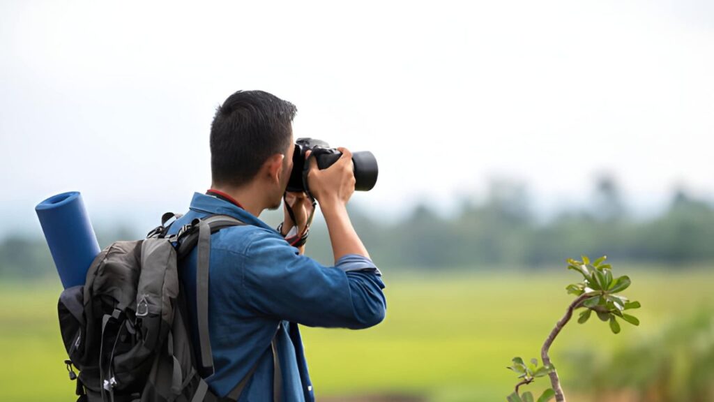 A person in a blue shirt and backpack takes a photo with a camera, capturing a lush, green landscape. The scene conveys a sense of adventure and exploration.