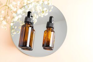 Amber glass dropper bottles on a round mirror with delicate white flowers. Minimalist setup on a soft peach background, conveying an elegant, serene vibe.
