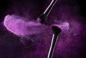 Two makeup brushes collide, releasing a cloud of vibrant purple powder against a dark background, creating a dynamic and artistic visual effect.