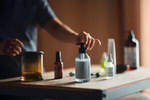 A person’s hand reaches for a soap dispenser on a wooden table with various skincare bottles and a jar, conveying a calm, organized atmosphere.