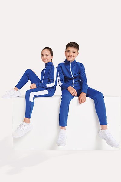 Two children in blue tracksuits sitting on a white box, smiling and looking at each other.
